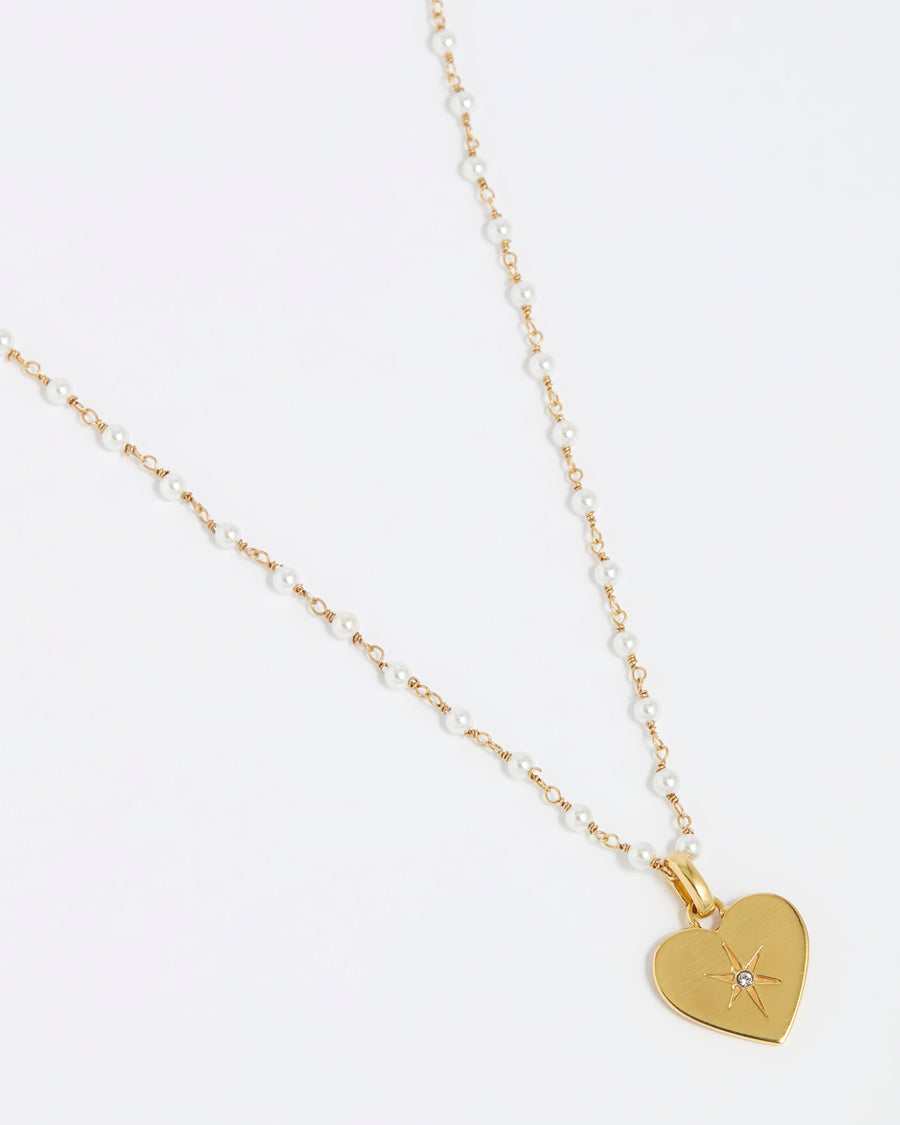 close up image shot of the Soru Jewellery clear crystal surrounded by a star etching set in a shiny gold heart attached to a white beaded necklace on a white background