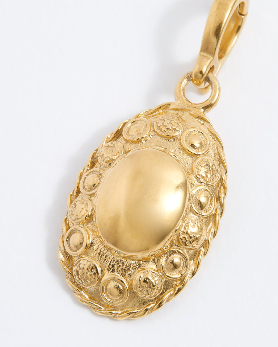 Close up of gold oval charm with intricate embossed patterns and a smooth central dome, featuring a small clasp for attachment.