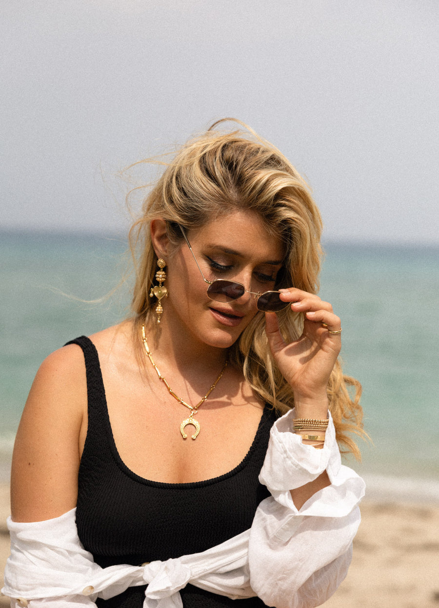 Daphne Oz wearing gold horseshoe charm and necklace outside at the beach looking down from her collaboration with soru 