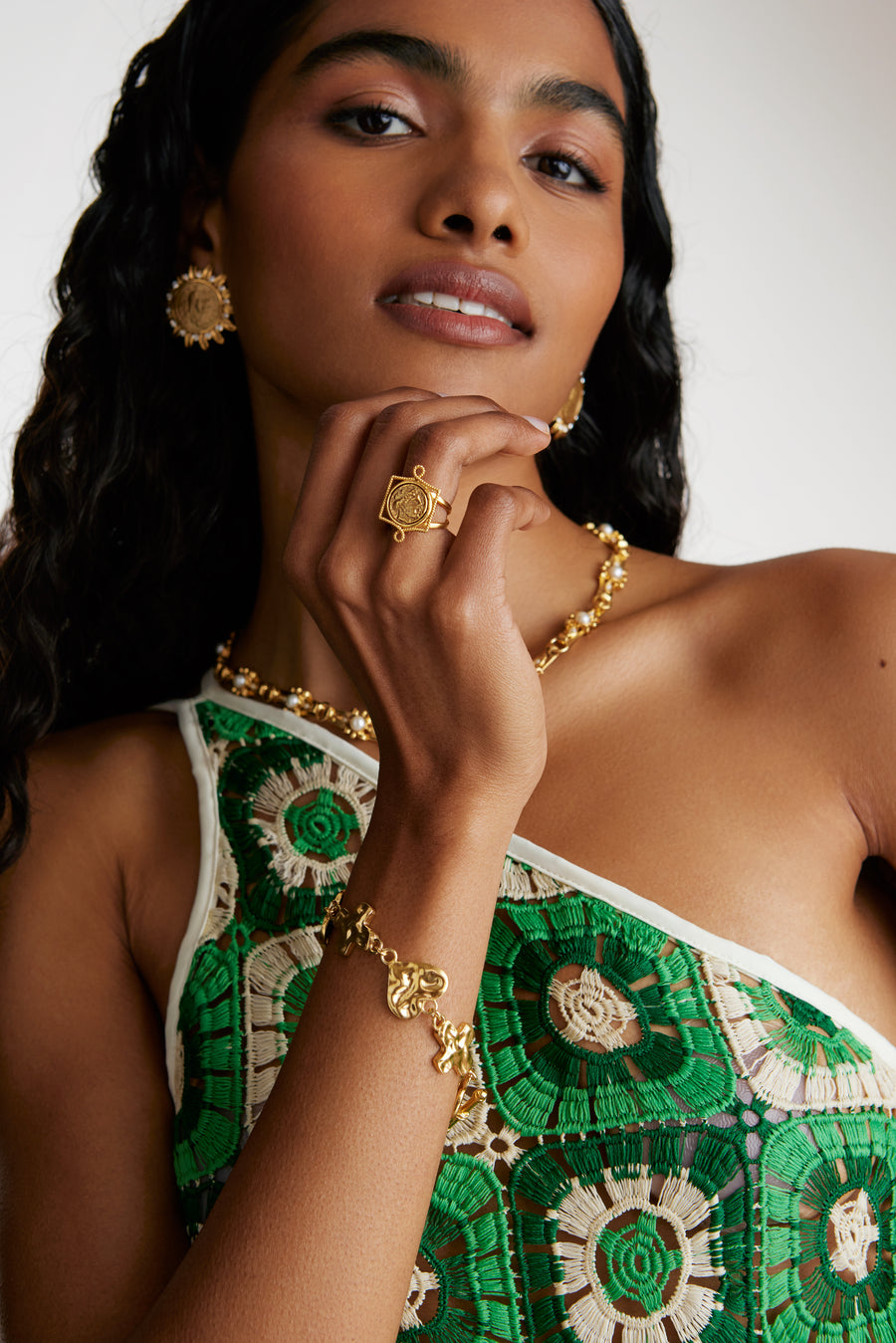 A model shot of dark haired model wearing soru jewellery medici coin ring, a coin shaped ring, with heart and cross bracelet, large coin earring and a green crocheted dress