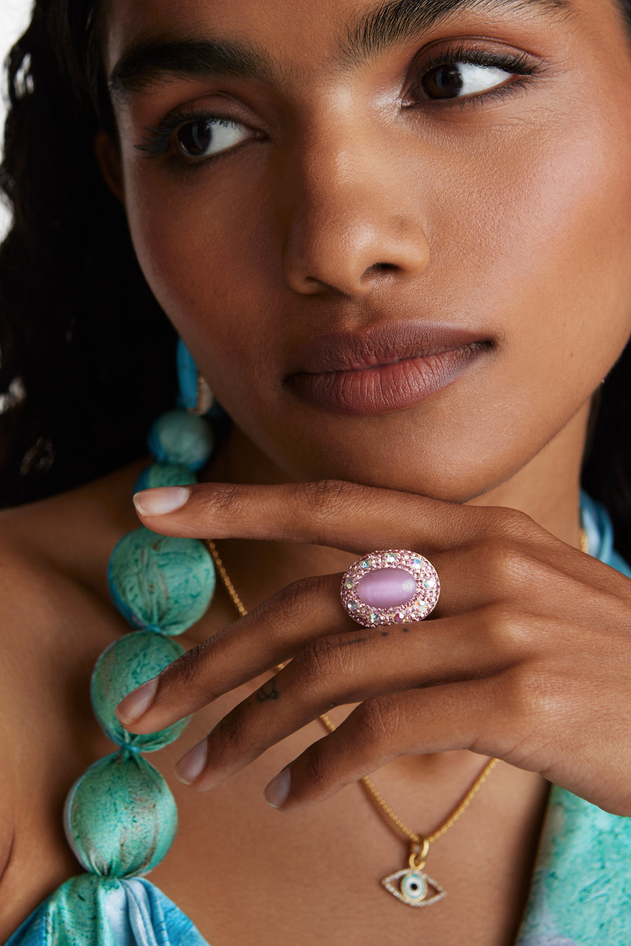 Model shot wearing chunky ring with pink centre stone and surrounded with crystals with hand under chin.  Model also wearing a delicate gold evil eye necklace