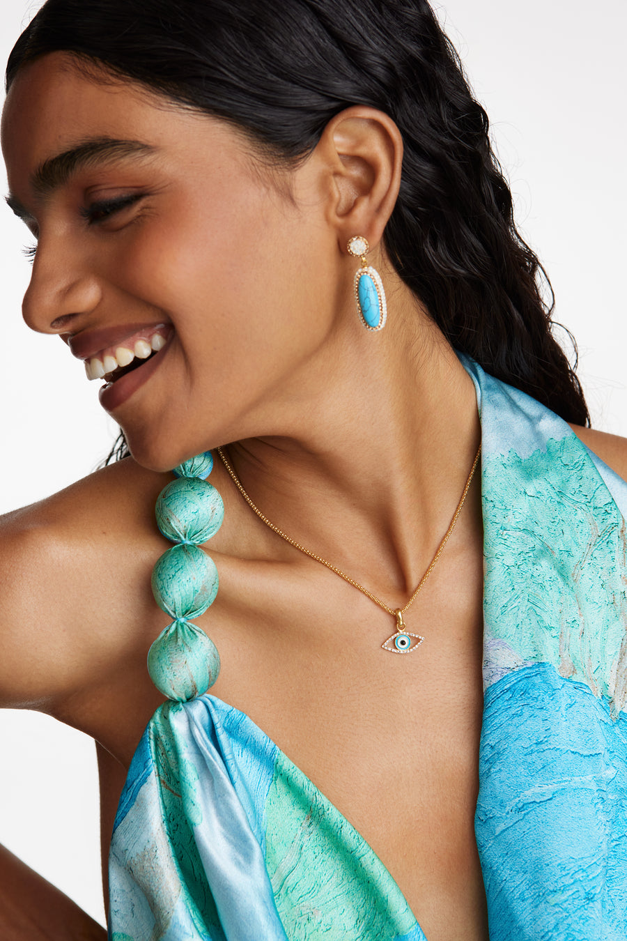 model shot wearing the crystal evil eye charm on a chain and also wearing turquoise drop earrings while smiling