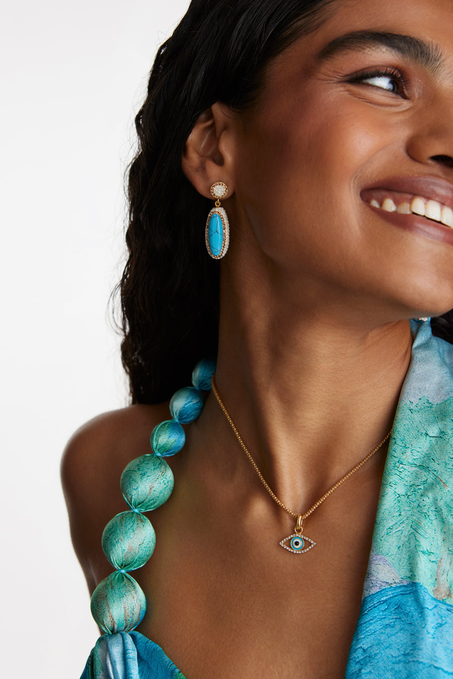 model shot wearing the crystal evil eye charm on a chain and also wearing turquoise drop earrings while smiling