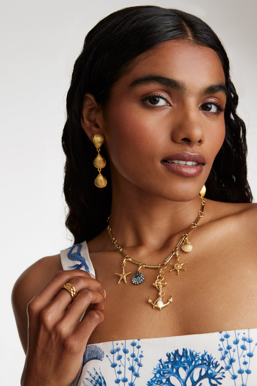 model shot wearing soru jewellery gold ships wheel charm with other nautical charms attached to a charm chain. Model also wearing shell drop earrings and a gold ring