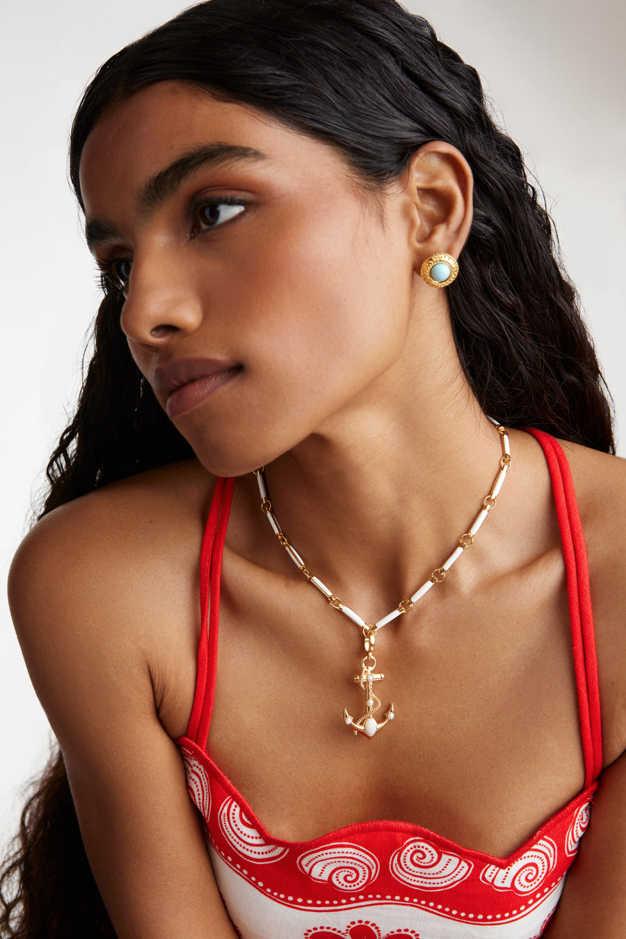 Model shot wearing the Soru Jewellery gold anchor charm with white embellishments and rope twist detail hung from a white and gold chunky charm chain. Model also wearing the round turquoise stud earrings while leaning forward