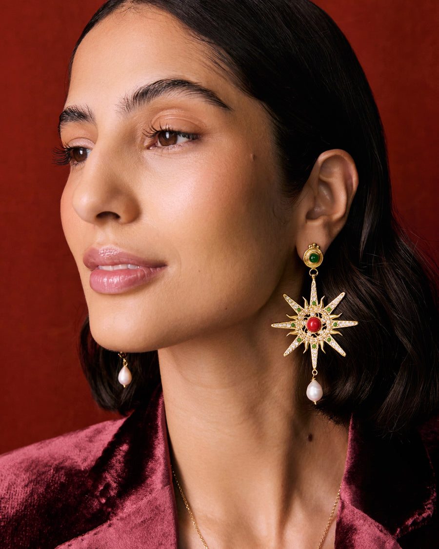 Model shot of lady wearing a mis-matched earring, one featuring a star. Detailed with hanging pearls and colourful green and red stones, on a red background.