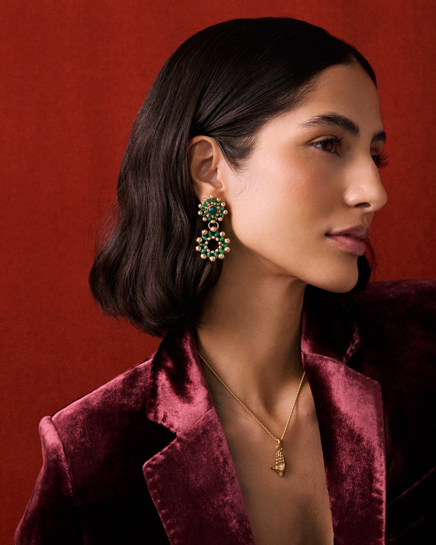 Model shot of lady wearing earrings featuring emerald green agate stones surrounded by delicate gold cutwork beading, in front of a red background. She's also wearing a charm necklace with a gold hand charm, as well as a red velvet blazer.