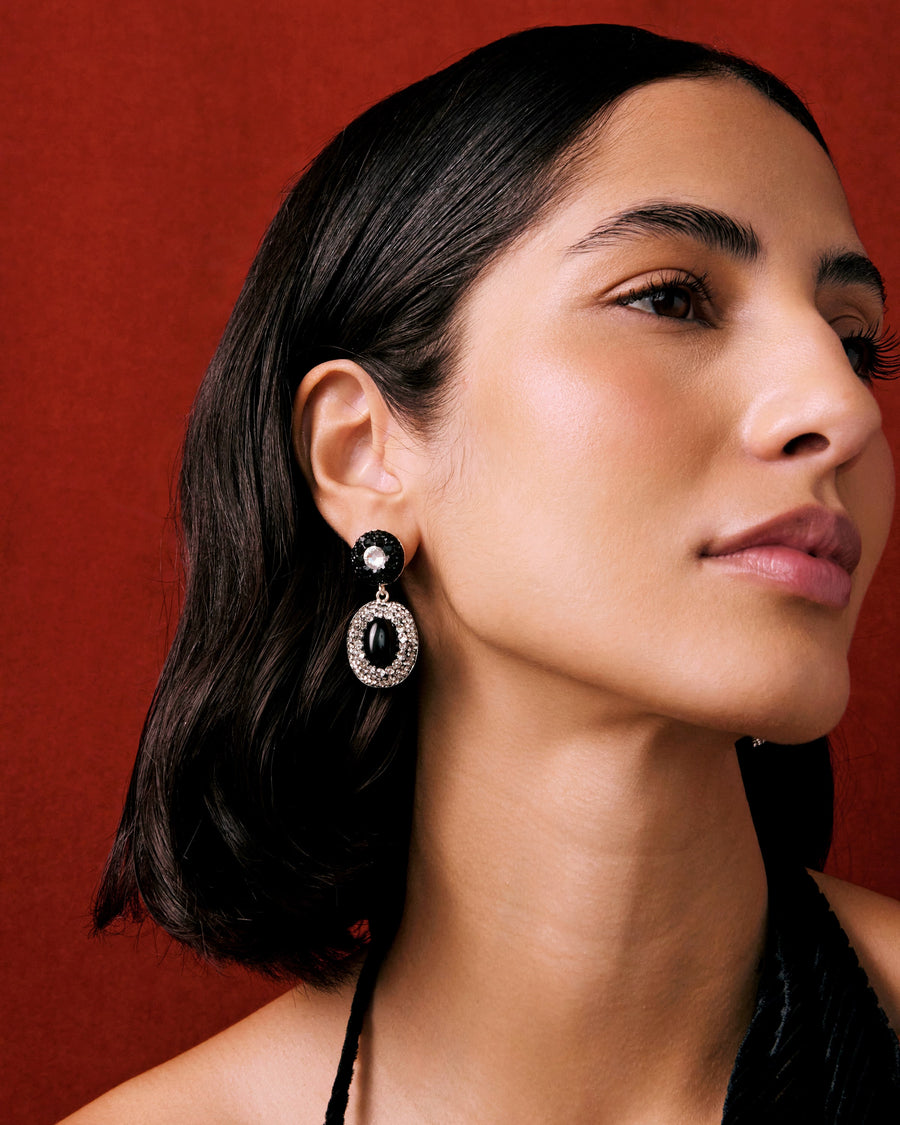 Model shot of lady wearing vintage style statement double drop earrings with a mix of glossy oval onyx gemstones with sparkling clear and black crystals, on a red background. 