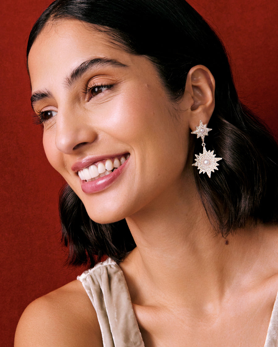 Model shot of lady wearing silver celestial earrings feature star burst shapes, encrusted with sparkling clear crystals and baroque pearl centres on a red background.