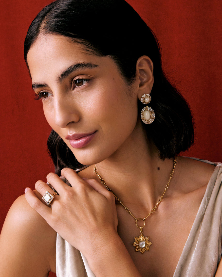 model wearing pearl earrings, flower shaped pearl necklace charm and a square pearl ring on a red background.