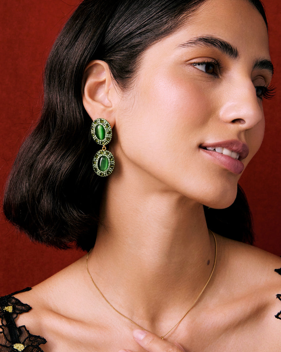 model shot of lady wearing double drop oval earrings featuring vibrant green cats eye stones, each surrounded by sparkling green crystals on a red background