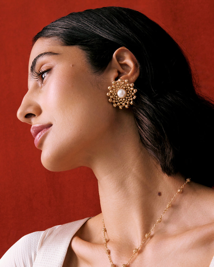 Model shot of a lady wearing a gold flower shaped beaded earring featuring a centre pearl gemstone on a red background.